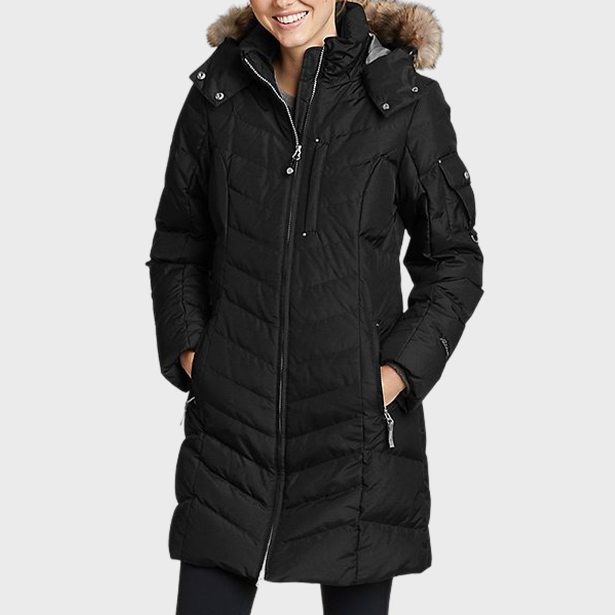 A person wears a long, black, puffer coat with hands in pockets against a plain, light background.