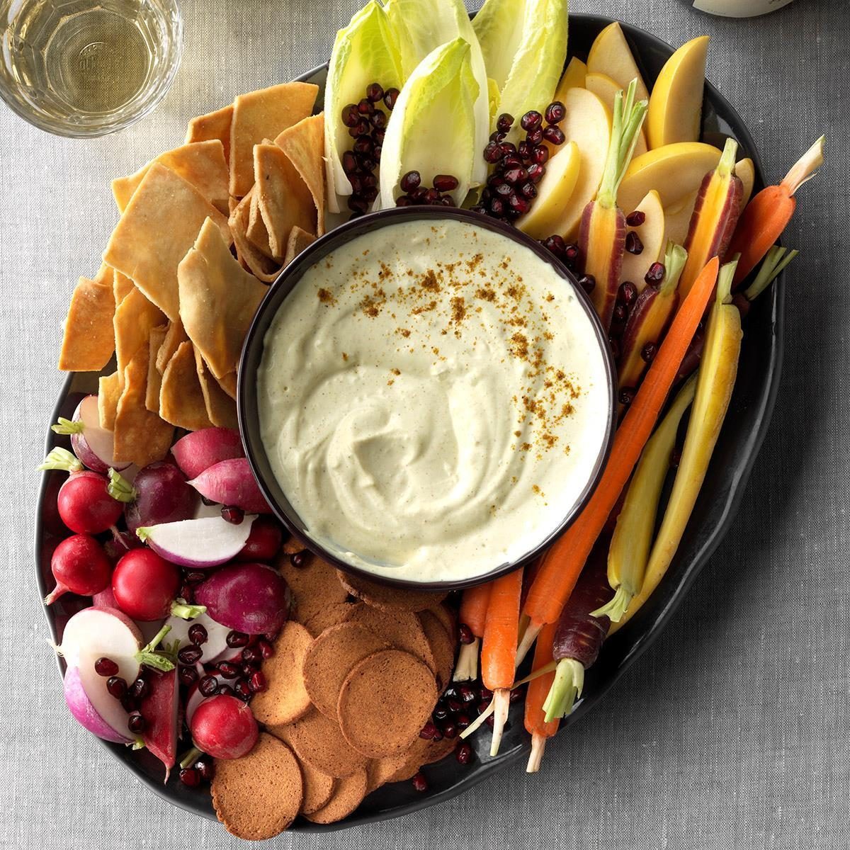 Dip sits in a bowl surrounded by fresh vegetables, crackers, and pomegranate seeds on a platter atop a gray tablecloth.