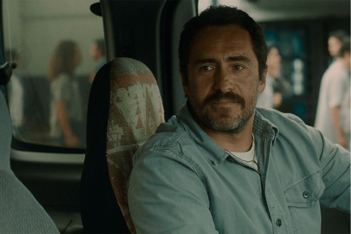 Man sits inside a vehicle, looking forward; wearing a light blue shirt in an urban environment with blurry figures outside.