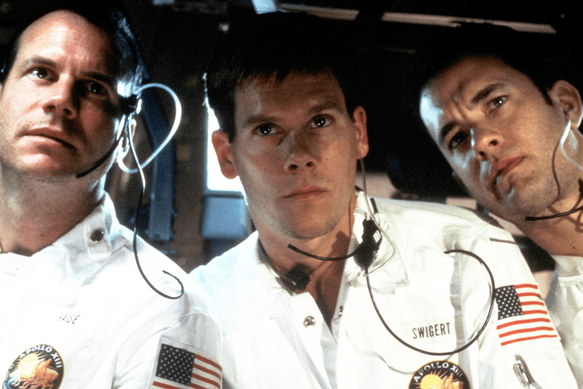 Three astronauts wearing headsets sit closely in a spacecraft, adorned with American and Apollo XIII patches, appearing focused and serious.