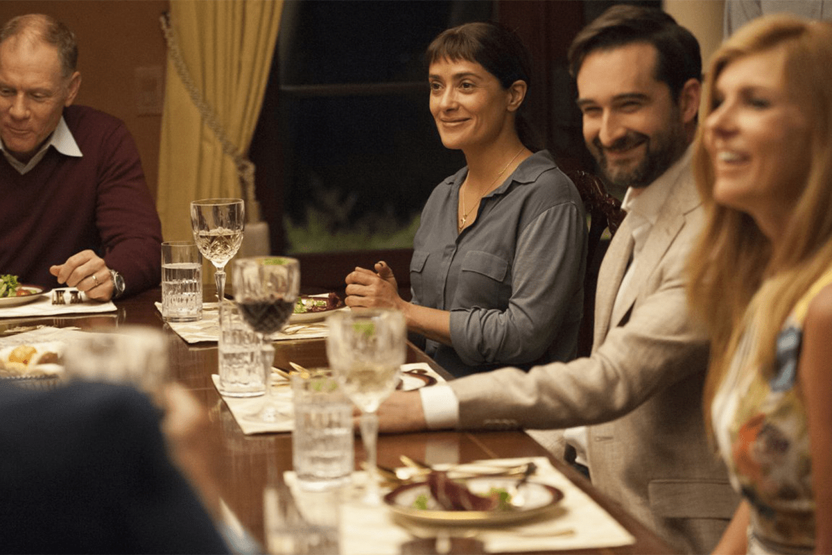 People dining, smiling, and conversing around a table set with wine glasses and plates of food, in a warmly lit room with curtains.