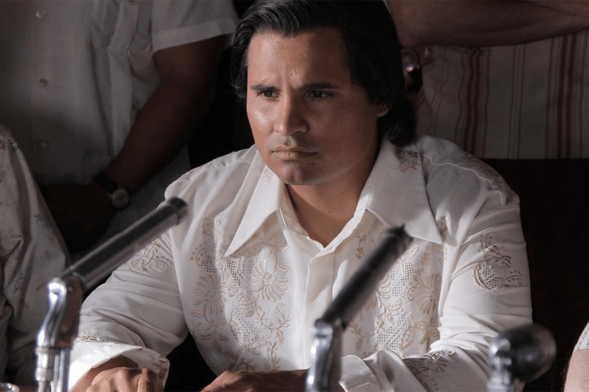 Person sits silently, facing microphones, wearing embroidered white shirt in a formal, indoor setting with others nearby.