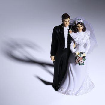 Plastic bride and groom figurine stands together, casting a shadow, on a white surface. Bride holds bouquet; groom wears a suit.