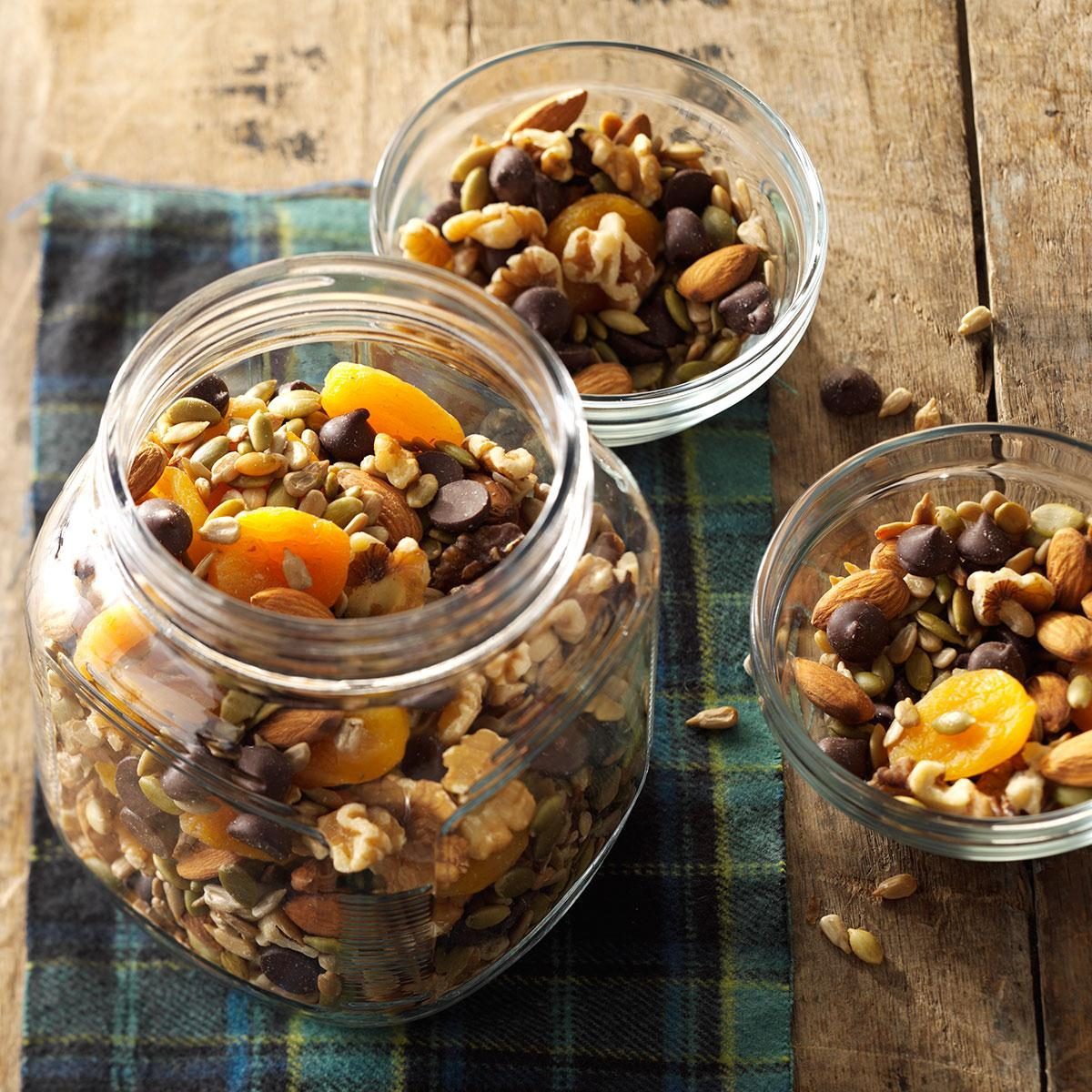 Trail mix sits in glass jars, containing nuts, seeds, dried apricots, and chocolate, resting on a wooden table covered with a plaid cloth.