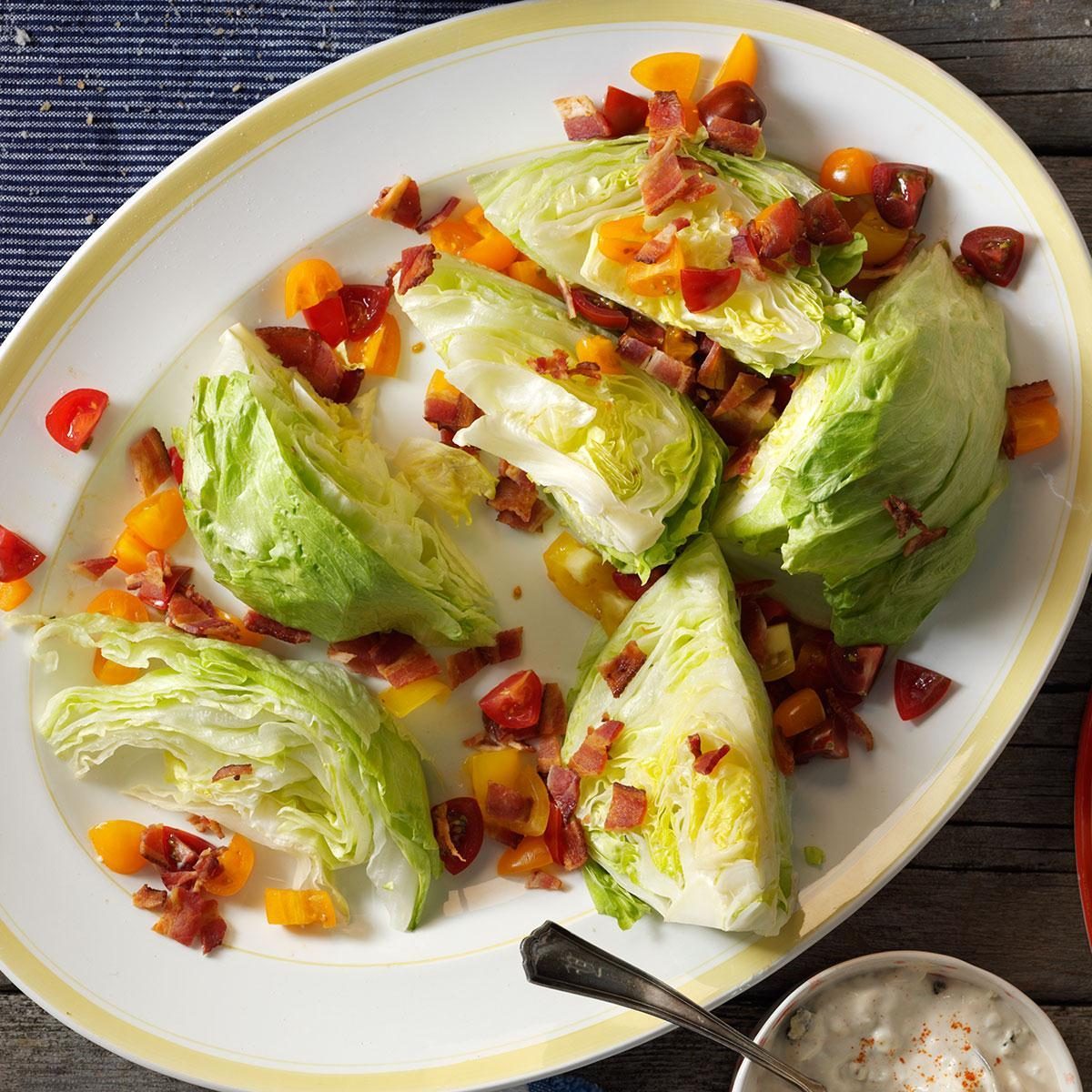 Wedge salad features iceberg lettuce topped with diced tomatoes and bacon on a white oval plate, surrounded by a rustic table setting.
