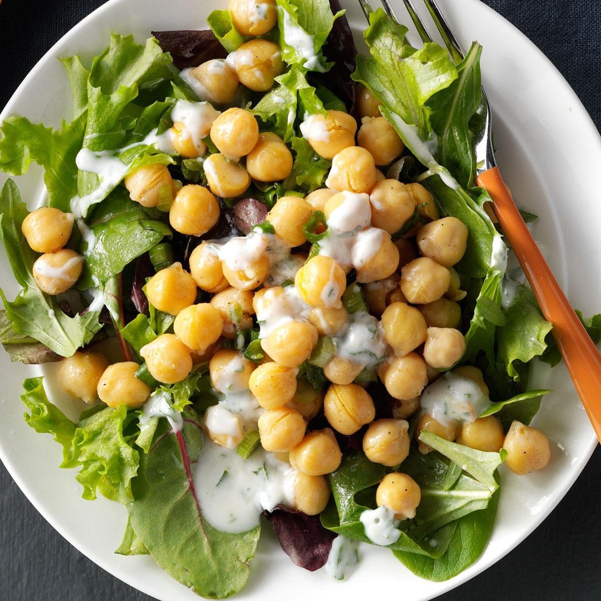 Chickpeas sit atop mixed greens, drizzled with creamy dressing, on a white plate with an orange-handled fork.