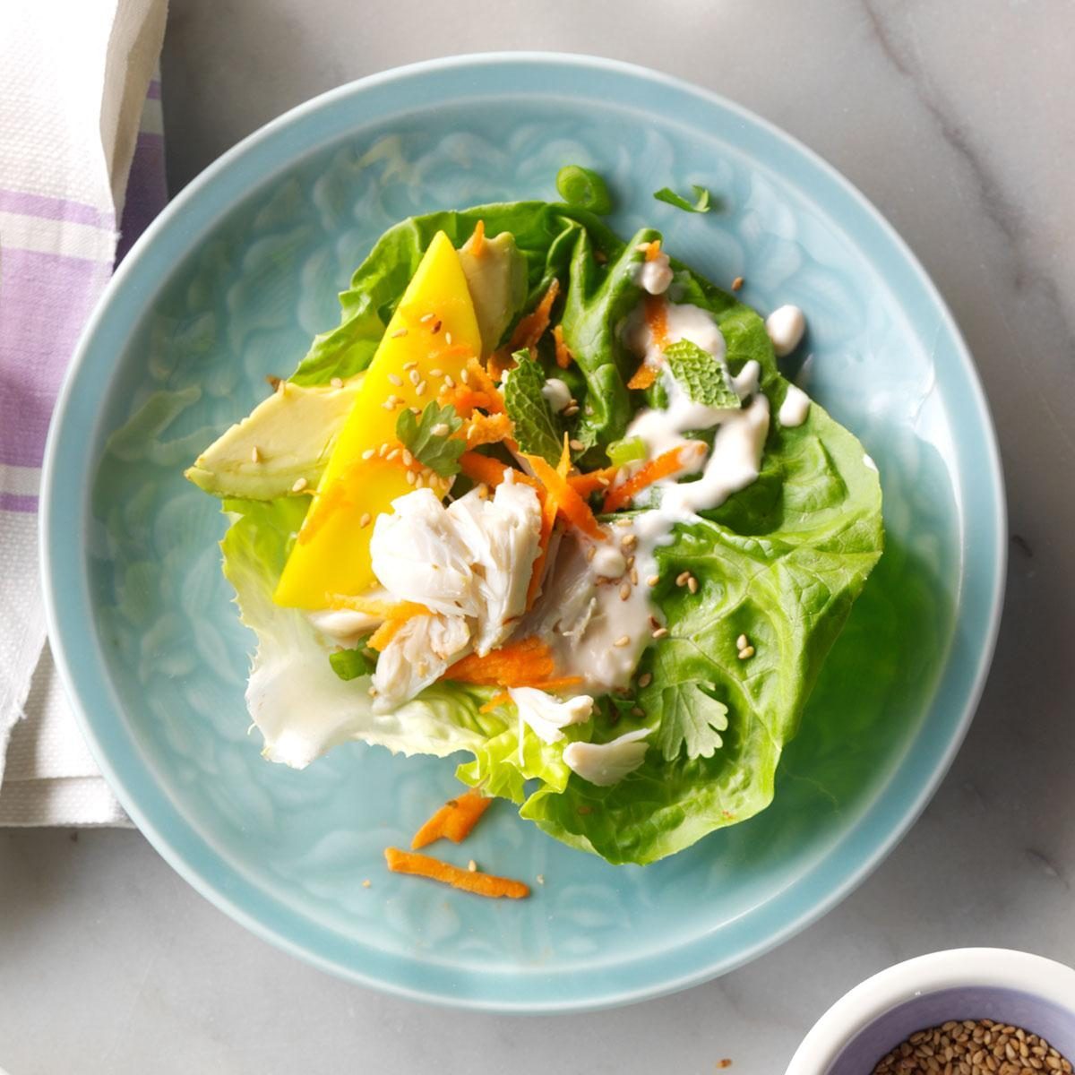 Lettuce wrap holds crab, mango, avocado, carrot, topped with sesame seeds and dressing on a blue plate beside a small sesame seeds bowl.