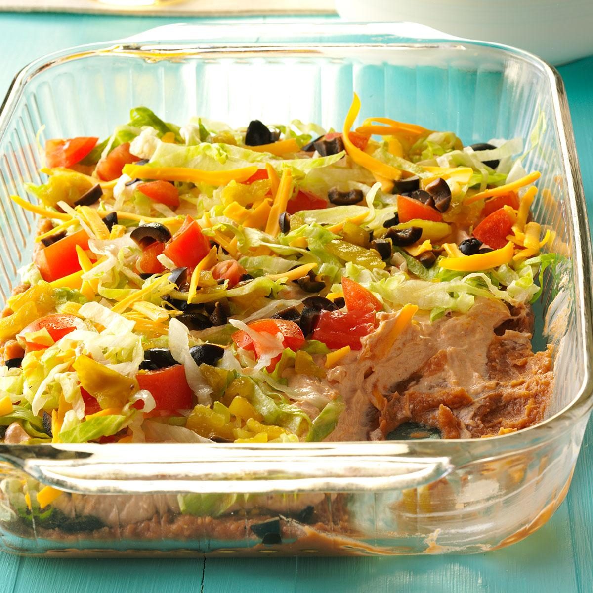 Glass dish holds layered dip with lettuce, tomatoes, olives, cheese, and refried beans; placed on a blue surface.