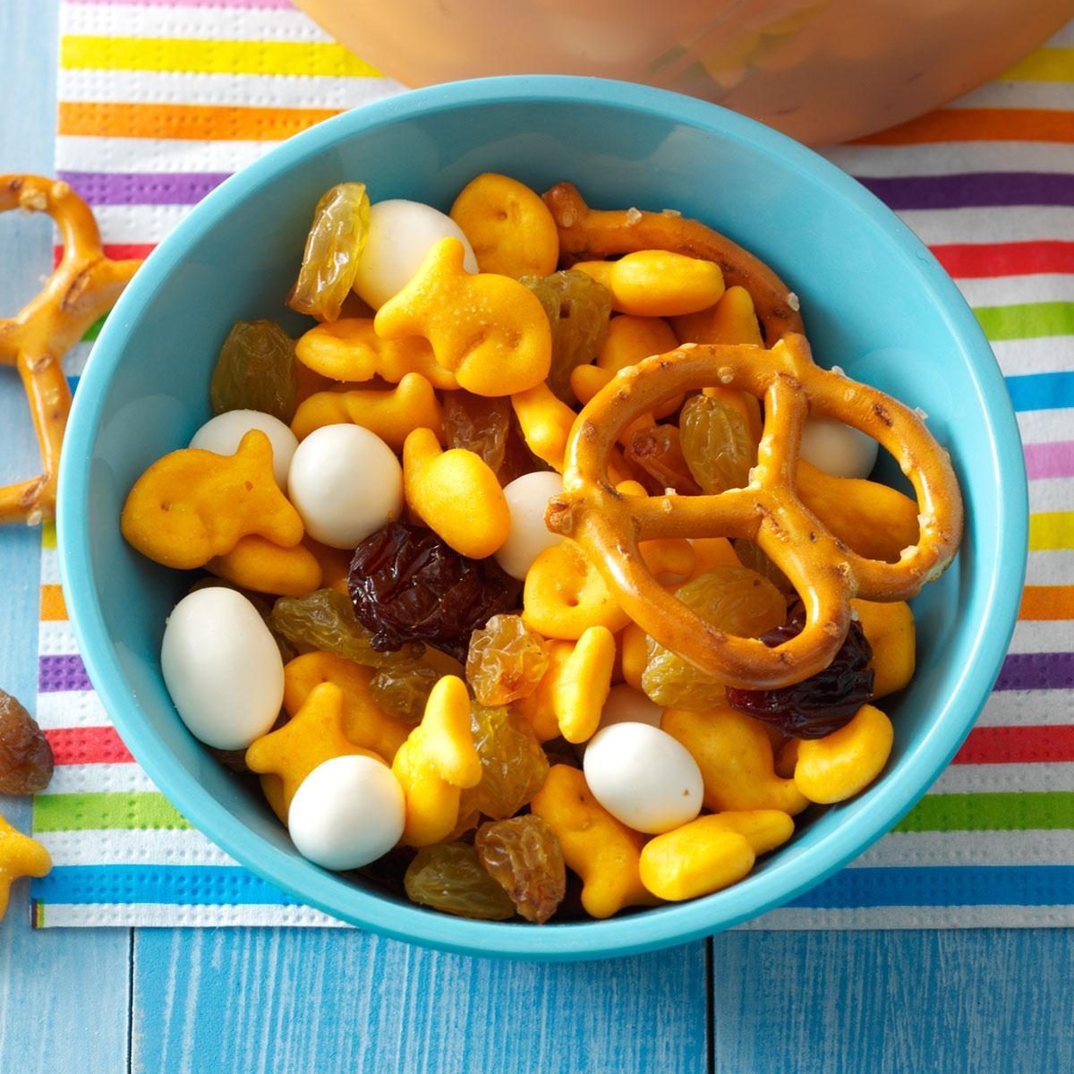 Snack mix fills a blue bowl, including fish-shaped crackers, pretzels, raisins, and yogurt-covered snacks on a colorful striped surface.