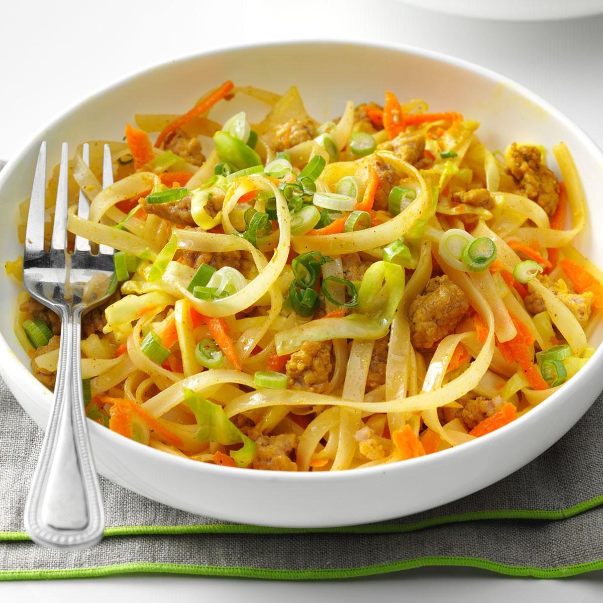 A white bowl holds stir-fried noodles with vegetables and meat, garnished with green onions, resting on a gray napkin with a fork.