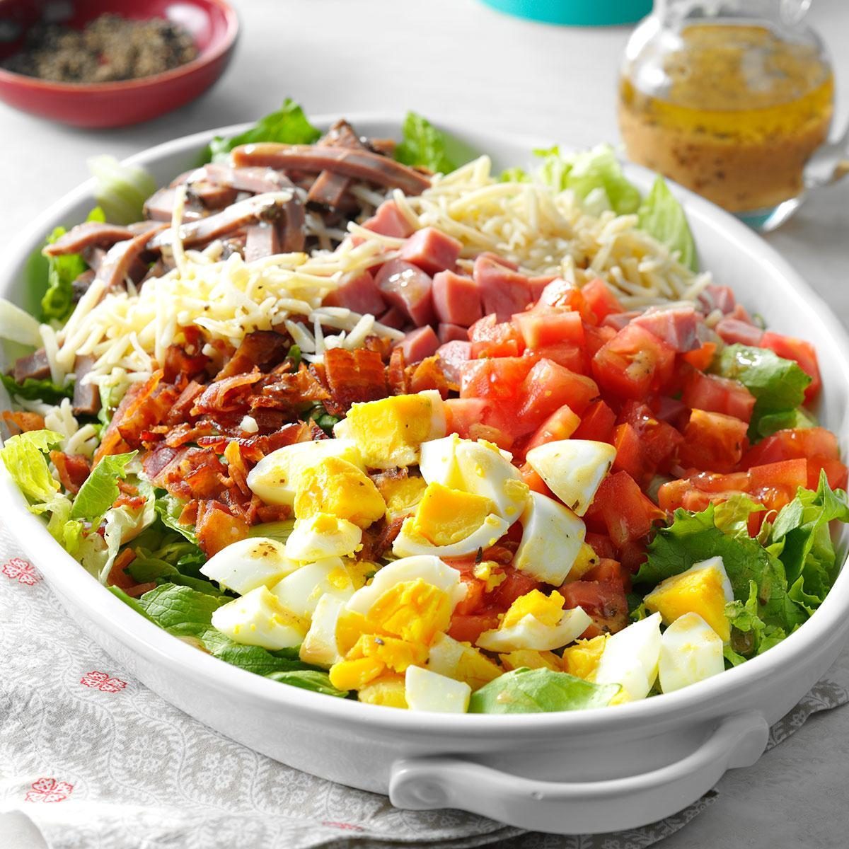 Salad features diced eggs, tomatoes, ham, bacon, cheese, and lettuce in a white dish, with dressing and pepper visible in the background.