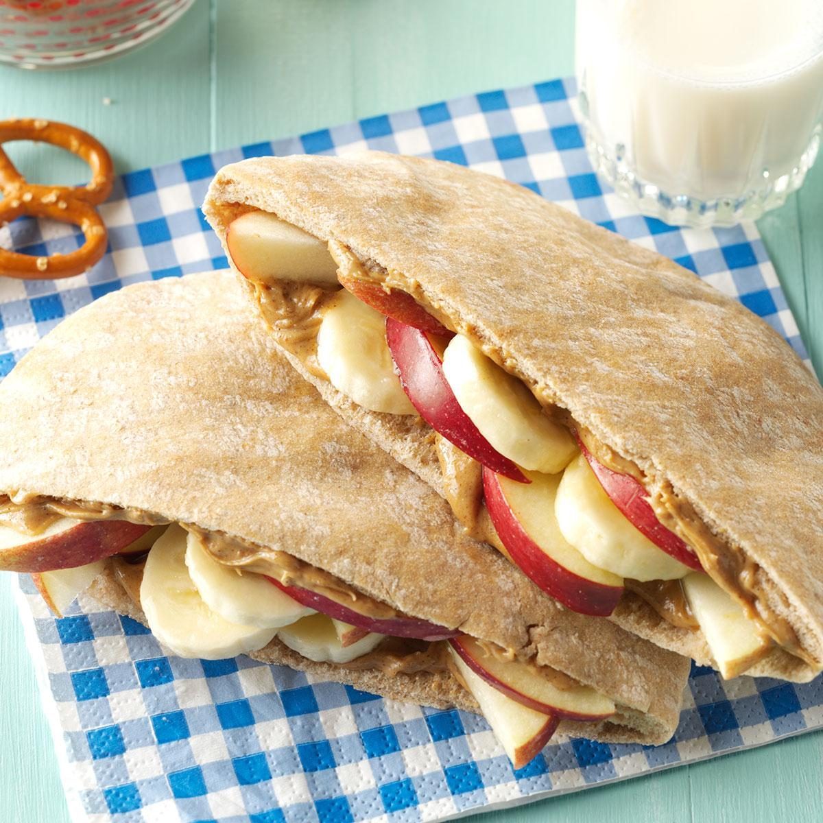 Pita bread pockets filled with peanut butter, apple, and banana slices rest on a blue-checkered napkin, next to a pretzel and glass of milk.