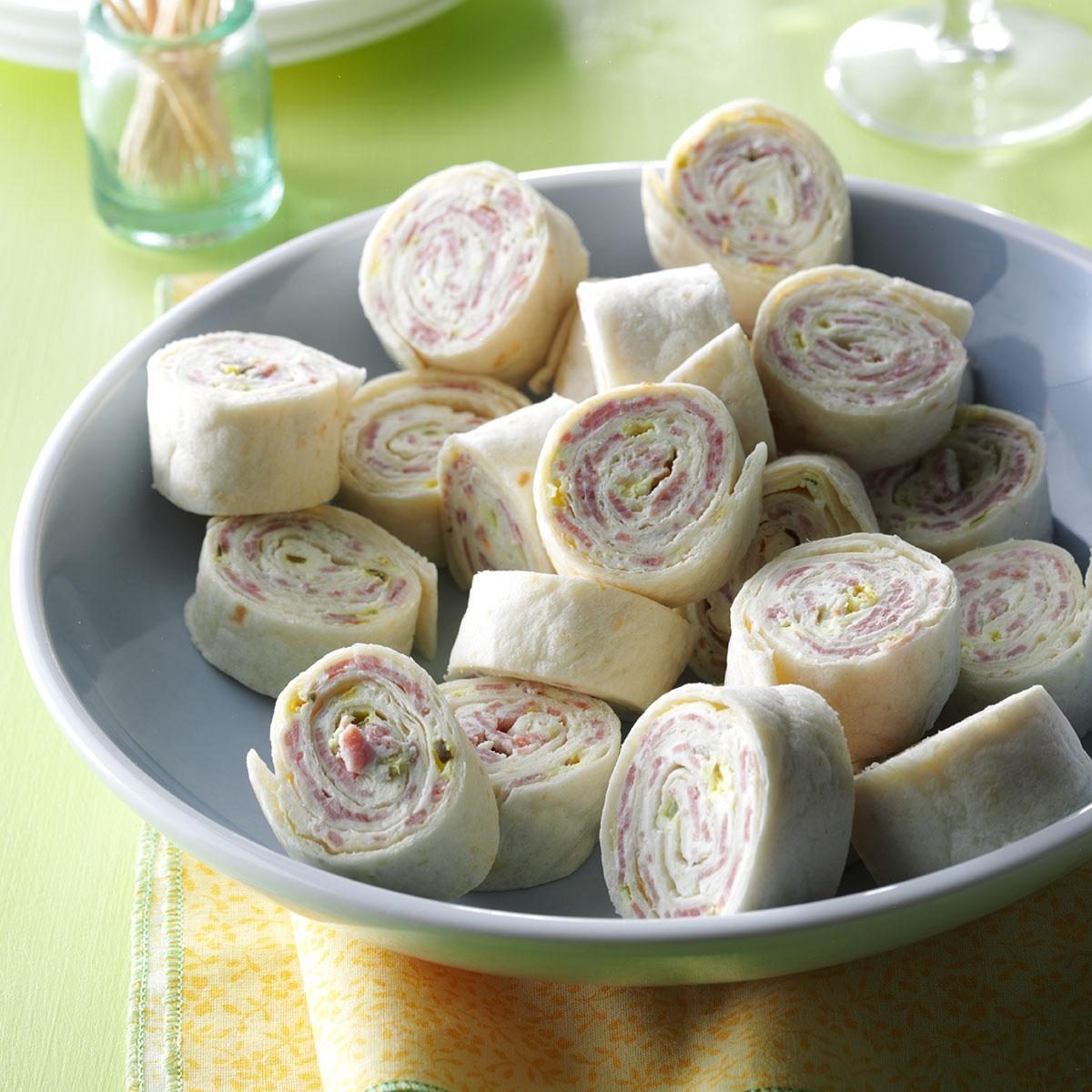 Pinwheel sandwiches lie in a bowl, displaying swirled layers of meat and spreads. A green table and yellow napkin complement the presentation.