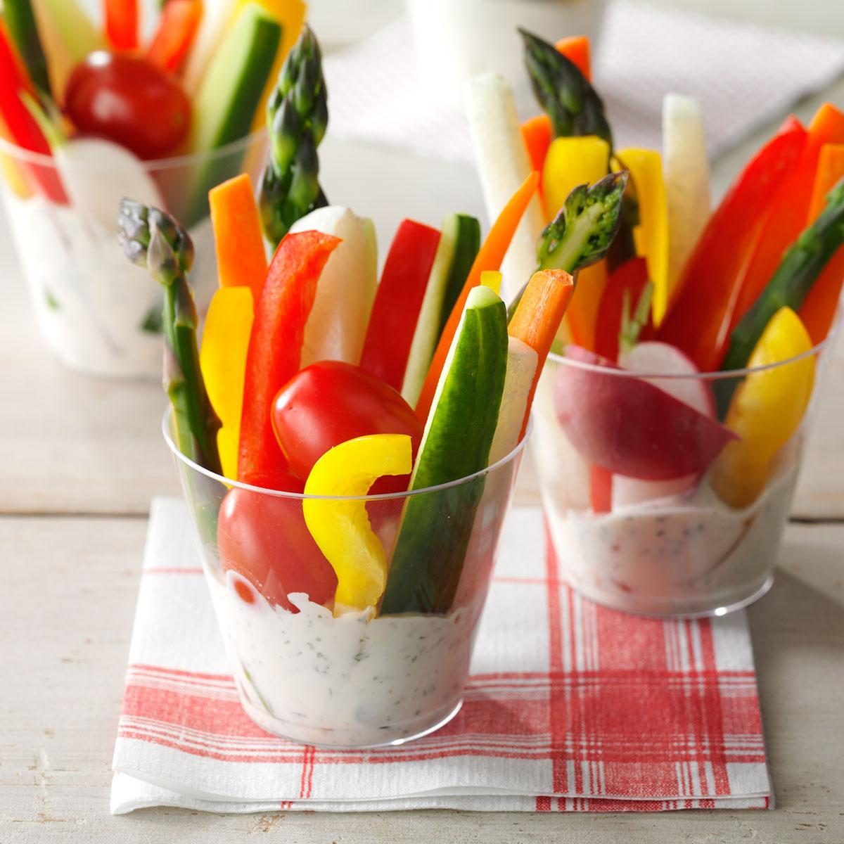 Vegetable sticks stand in clear cups filled with creamy dip, placed on red plaid napkins on a light wooden surface.