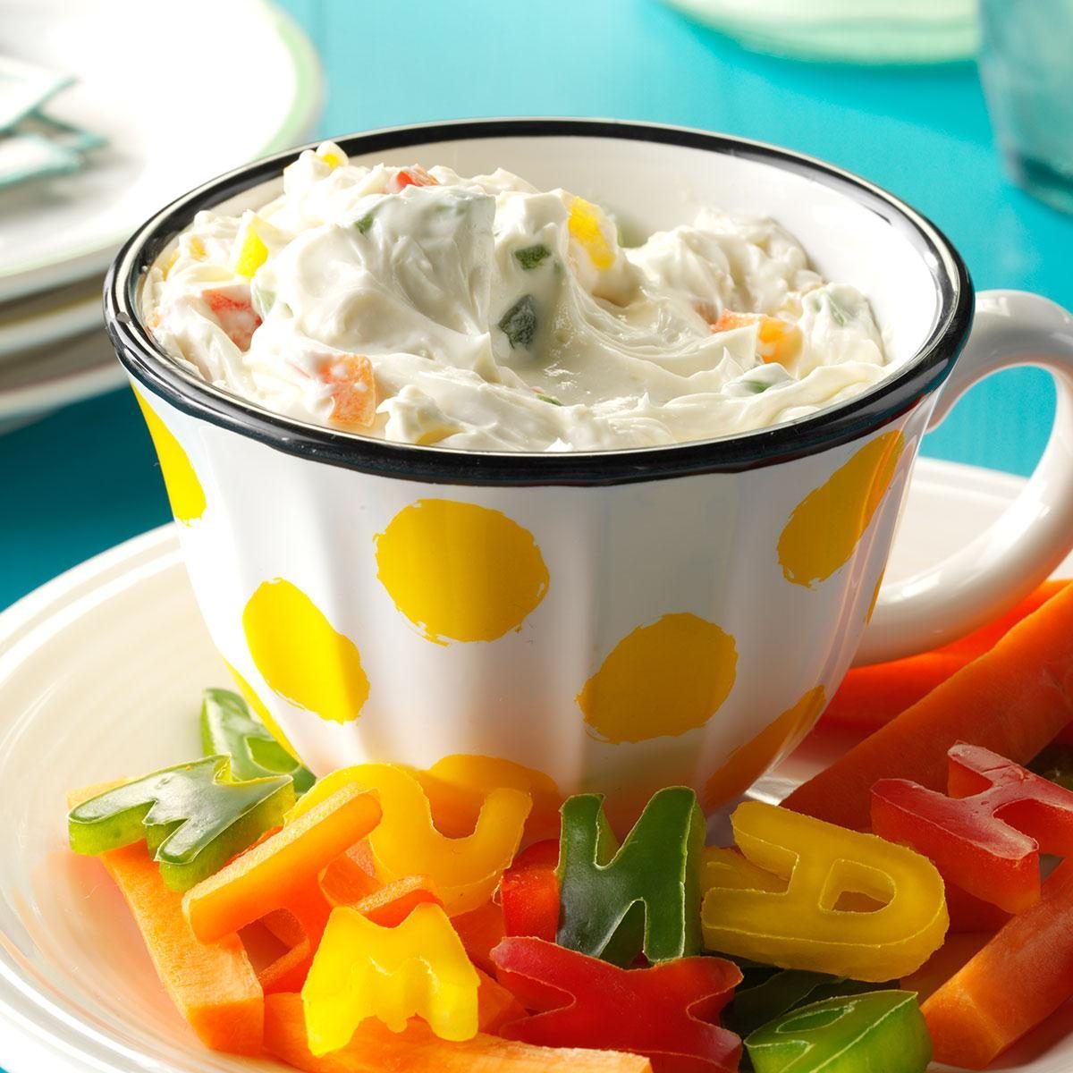 Cup holding creamy dip sits on a plate, surrounded by colorful vegetable shapes and carrot sticks, with a bright blue background.