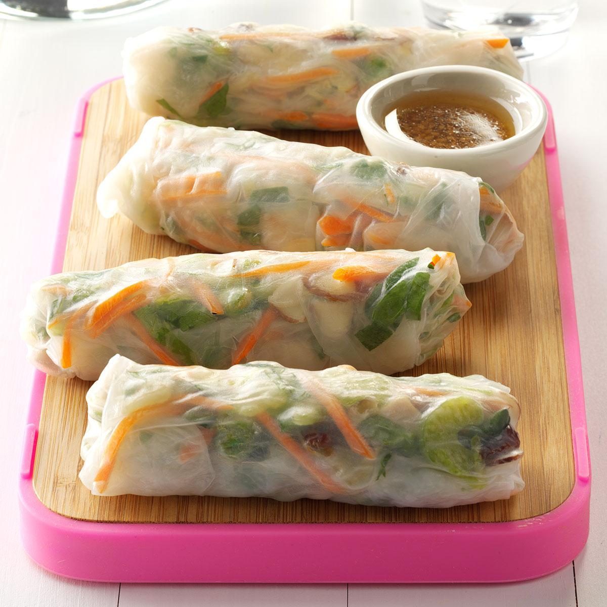 Spring rolls rest on a wooden tray, accompanied by a small bowl of dipping sauce, set in a bright, minimalist environment.