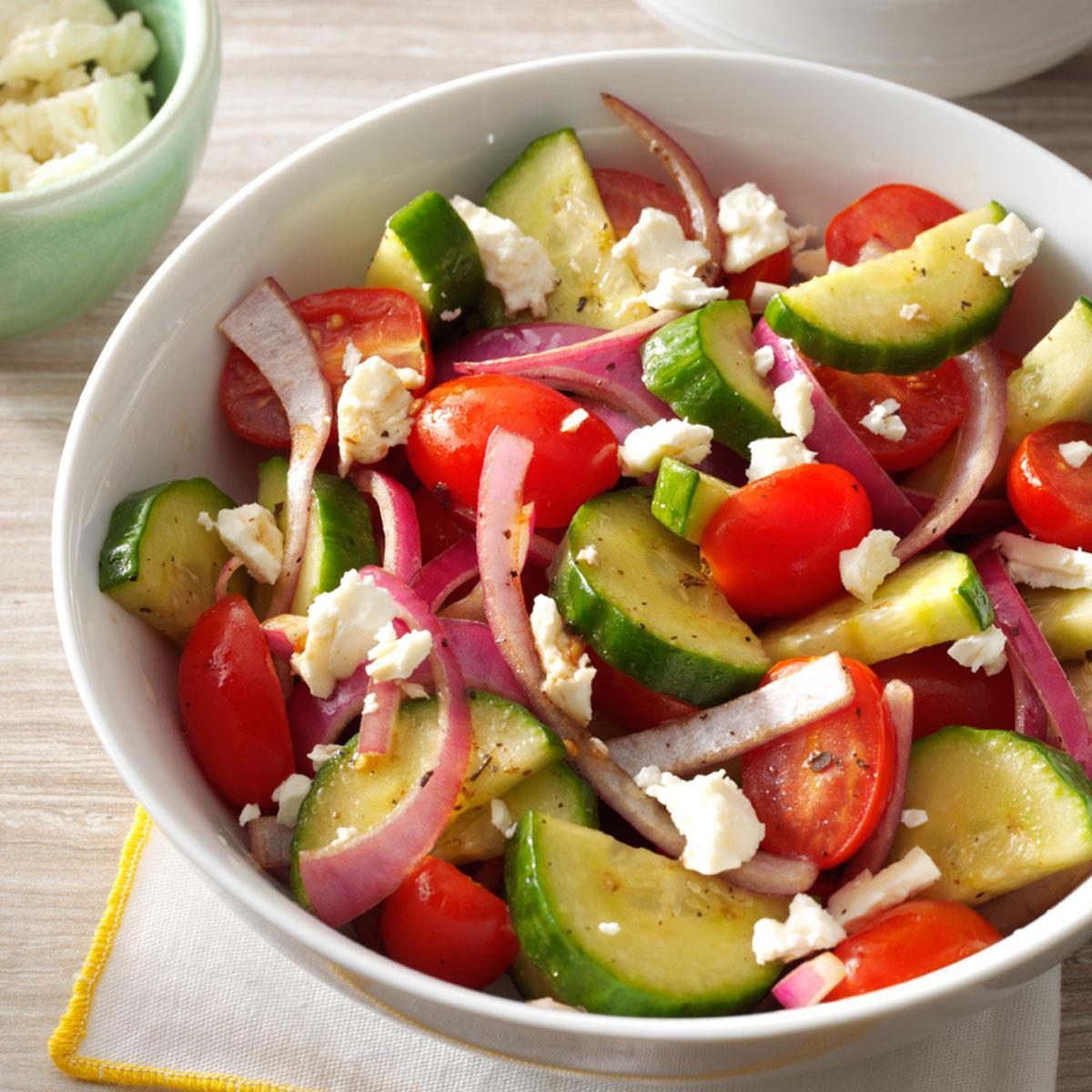 A bowl contains a fresh salad with cucumber slices, cherry tomatoes, red onion, and feta cheese on a light wooden table.