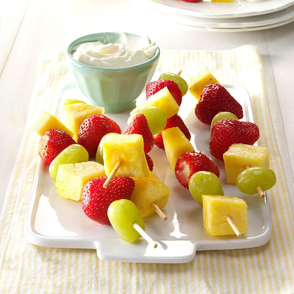 Fruit skewers rest on a white tray, consisting of strawberries, pineapple, and grapes, accompanied by a small bowl of creamy dip nearby.