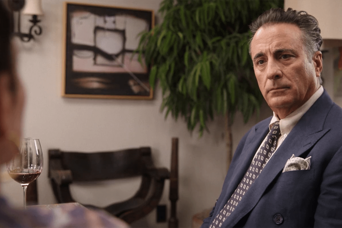 A man in a blue suit sits attentively indoors, with abstract art, a plant, and a glass of wine visible.