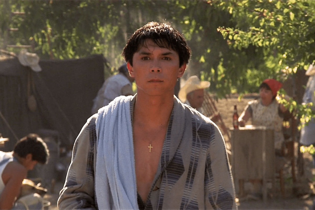 Young person stands draped in a towel, wearing a cross necklace, in a sunlit village scene with people and trees in the background.