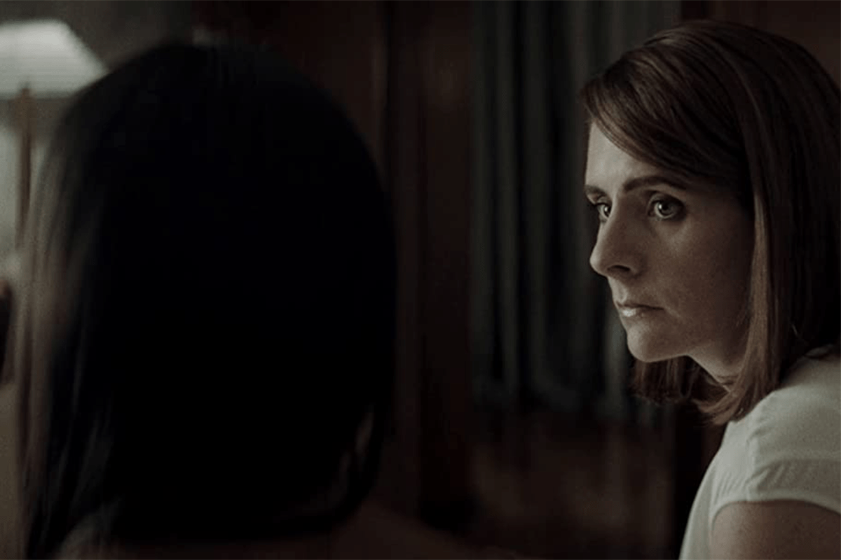 A woman looks intently at another person in a dimly lit room, with curtains in the background.