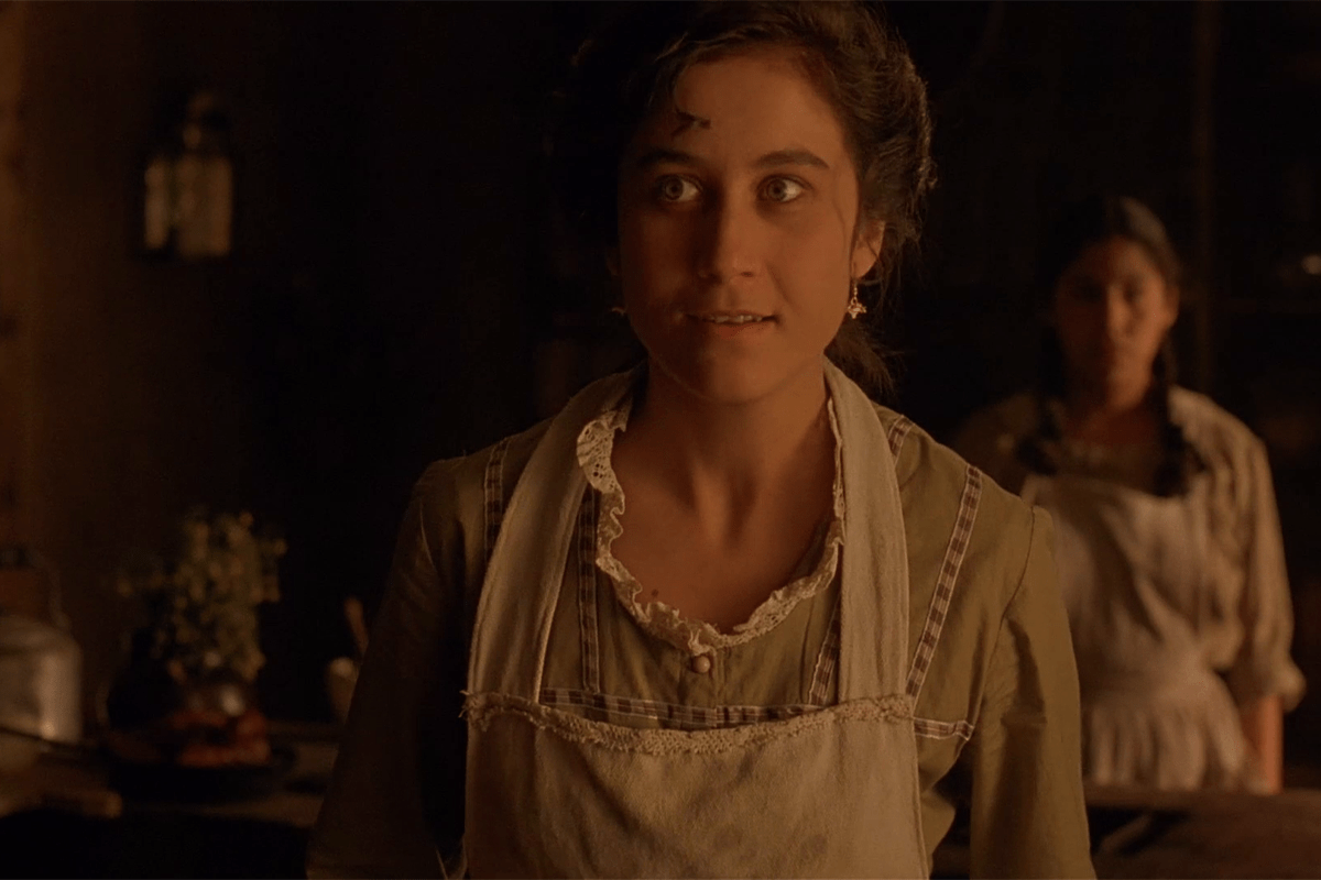 Young woman stands in dimly lit kitchen, wearing a vintage apron, with another woman blurred in the background.