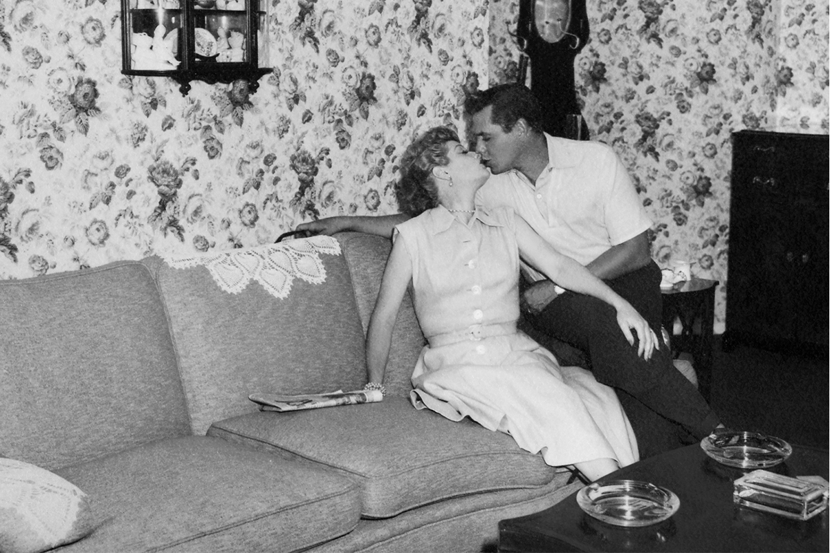 A couple sitting closely kisses on a sofa in a living room with floral wallpaper and decorative figurines on a wall shelf.