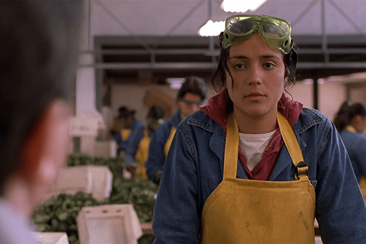 Woman wearing safety goggles and yellow apron works in a factory setting, surrounded by other workers in similar attire and equipment.