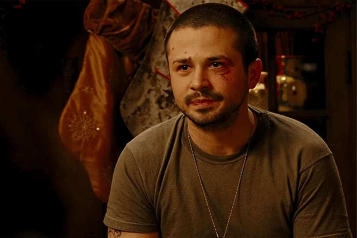 A man sits calmly with a neutral expression, wearing a gray t-shirt and chain necklace, indoors with warm lighting and decorative items behind.