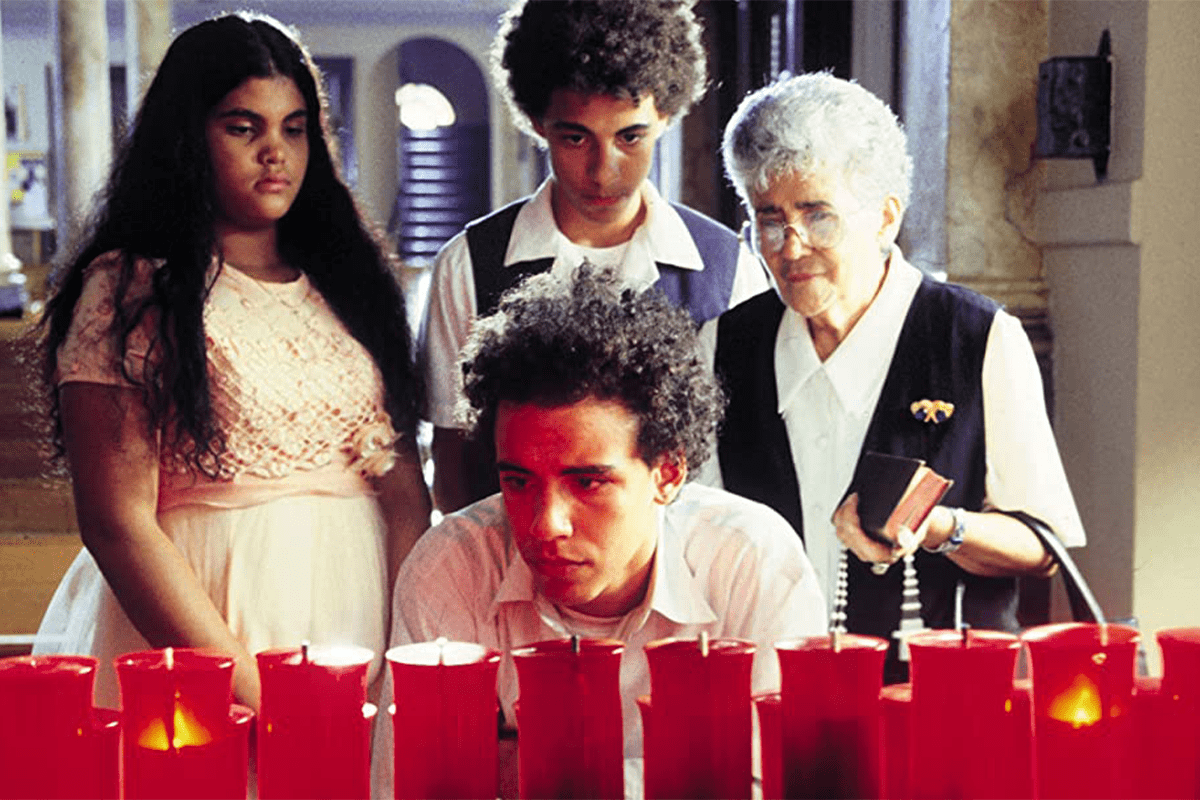 Four people gaze silently at a row of red votive candles inside a dimly lit room with arched doorways.
