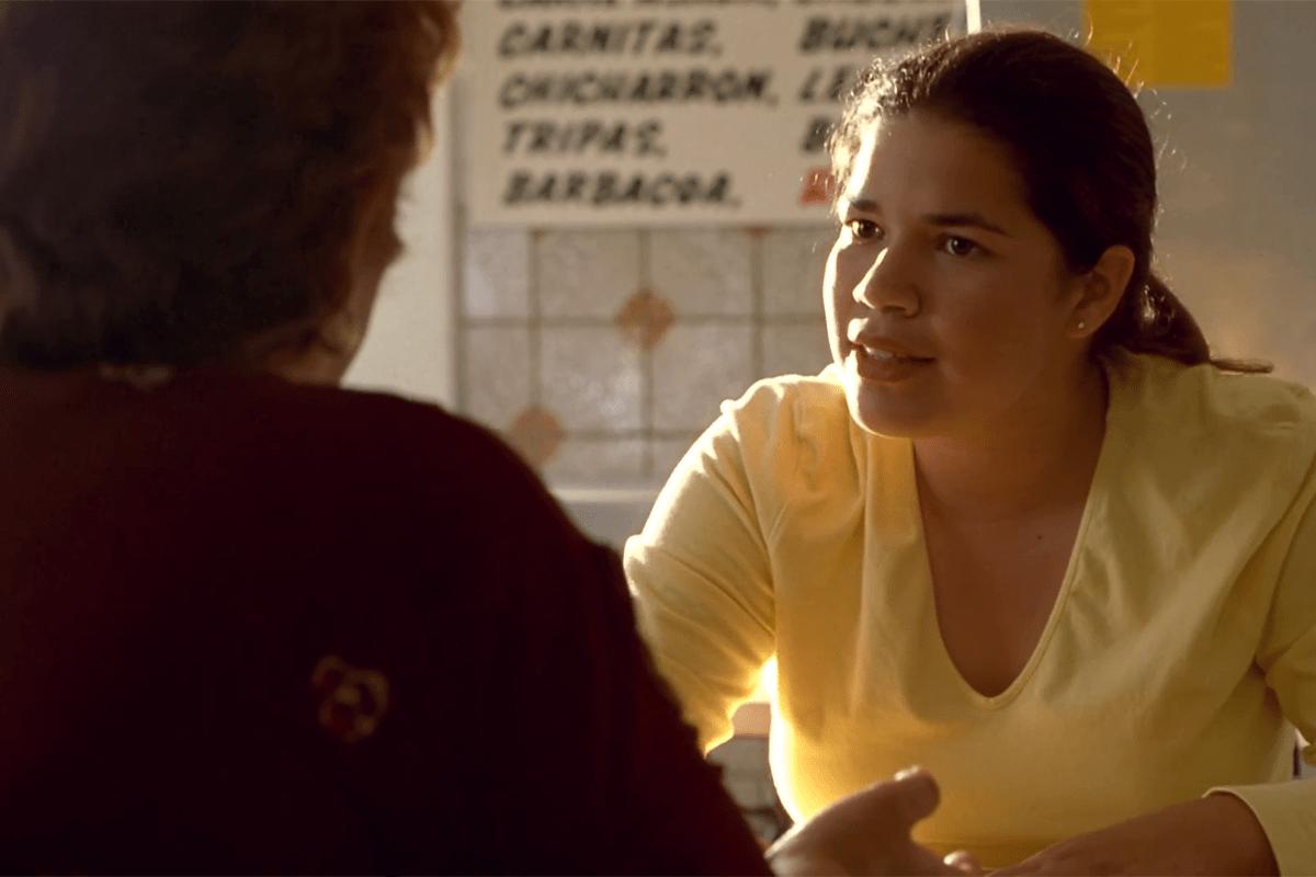 A woman, wearing a yellow shirt, converses intently with another person against a restaurant-like backdrop. A menu lists "Carnitas, ChicharrÃ³n, Tripas, Barbacoa."