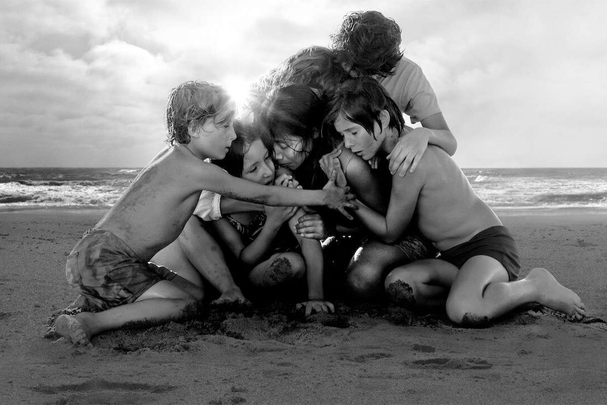 Children huddle together, embracing closely on a sandy beach with ocean waves behind them, creating a scene of companionship and warmth.