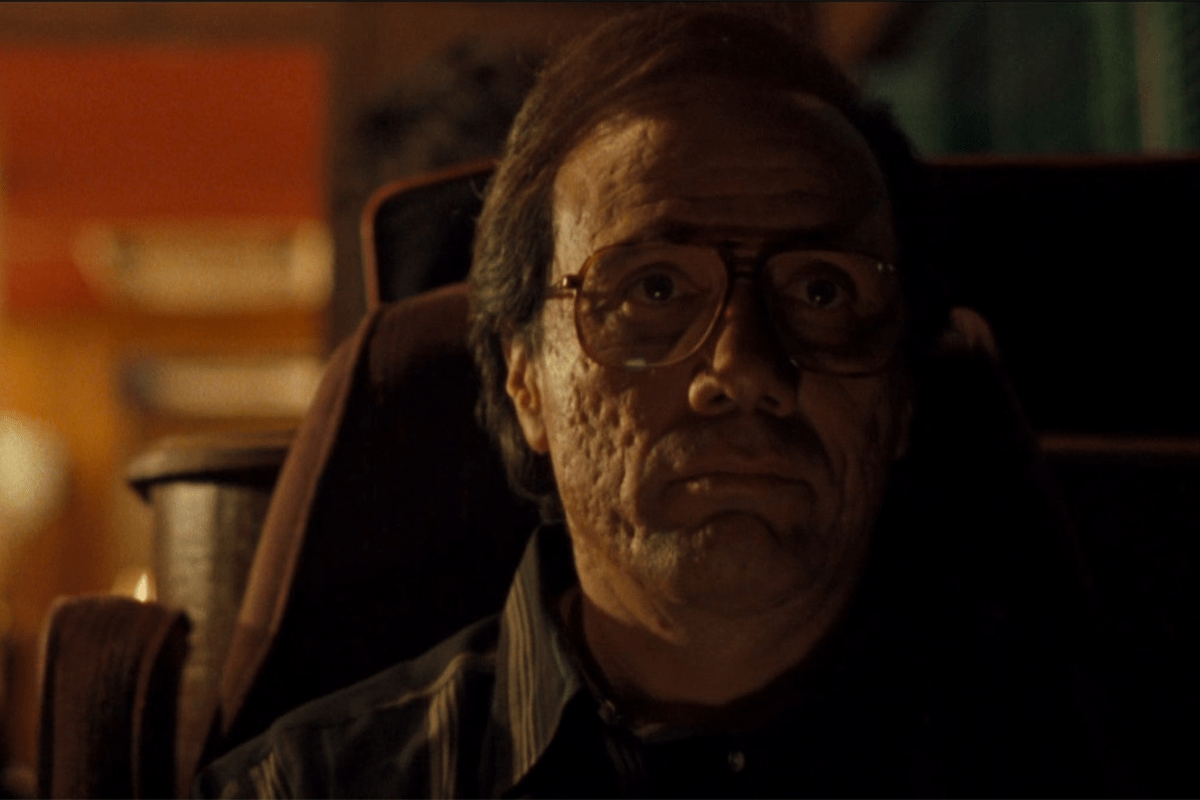 A person with glasses sits in low light, focused, surrounded by blurred, warm-colored furnishings.