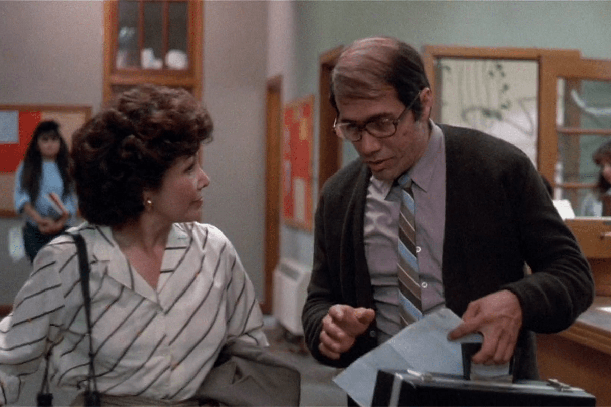 Two people converse in an office, one holding papers. A bulletin board and a person carrying books are visible in the background.