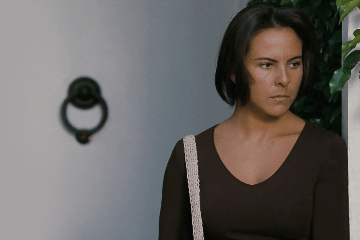 Woman stands pensively, wearing a dark top and crochet strap bag, near green leaves and a wall-mounted door knocker.