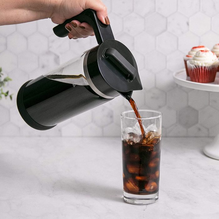Carafe pours dark liquid into a glass with ice on a marble counter, near cupcakes and a hexagonal-tiled wall.