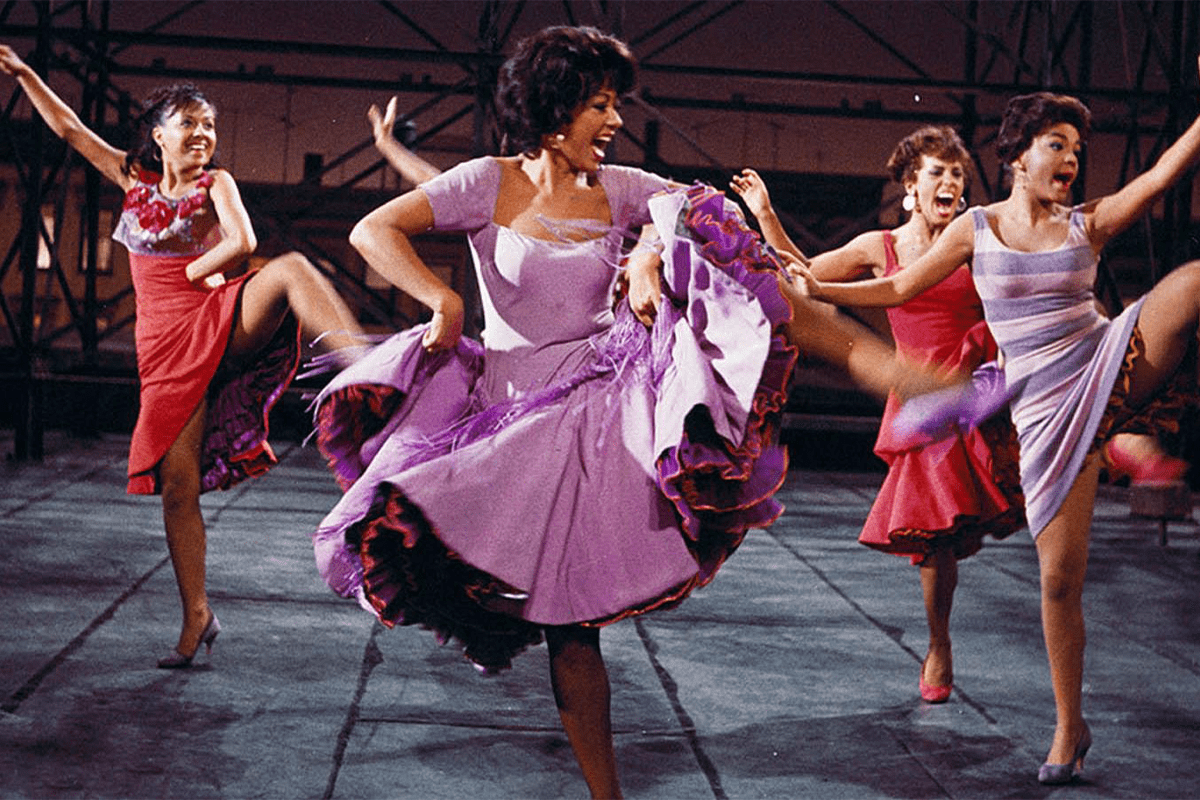 Four dancers energetically perform, kicking high, wearing brightly colored dresses in an indoor theatrical setting.
