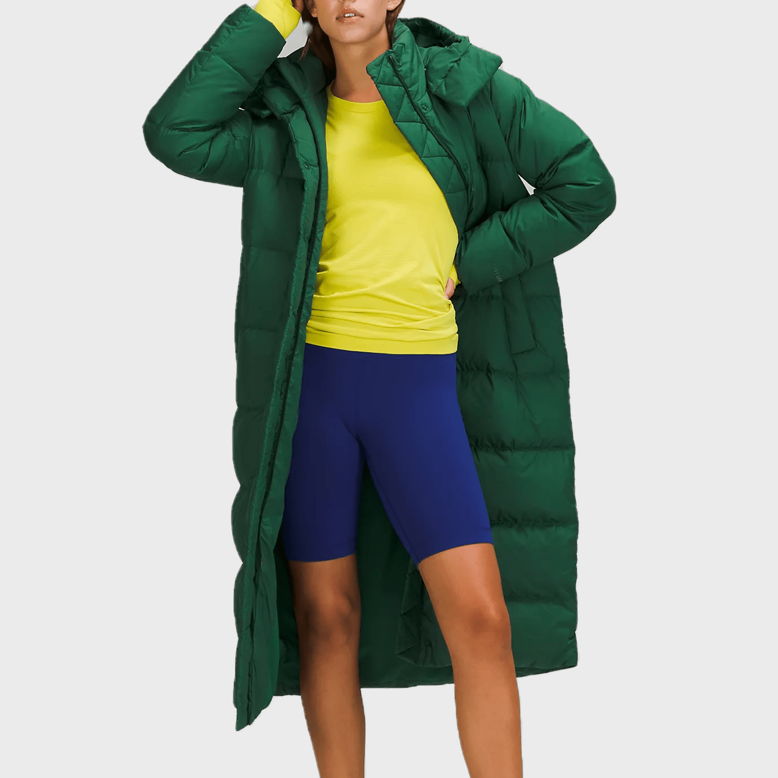 Person wearing a green puffy coat over a yellow shirt and blue shorts, standing in front of a plain, light gray background.