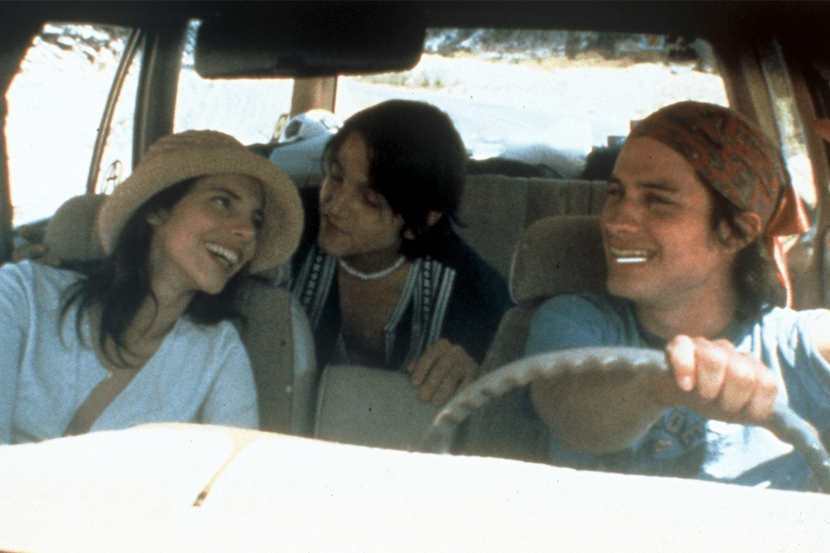 Three people smiling inside a car; the driver is steering, while two passengers converse happily in the backseat during a daytime road trip.