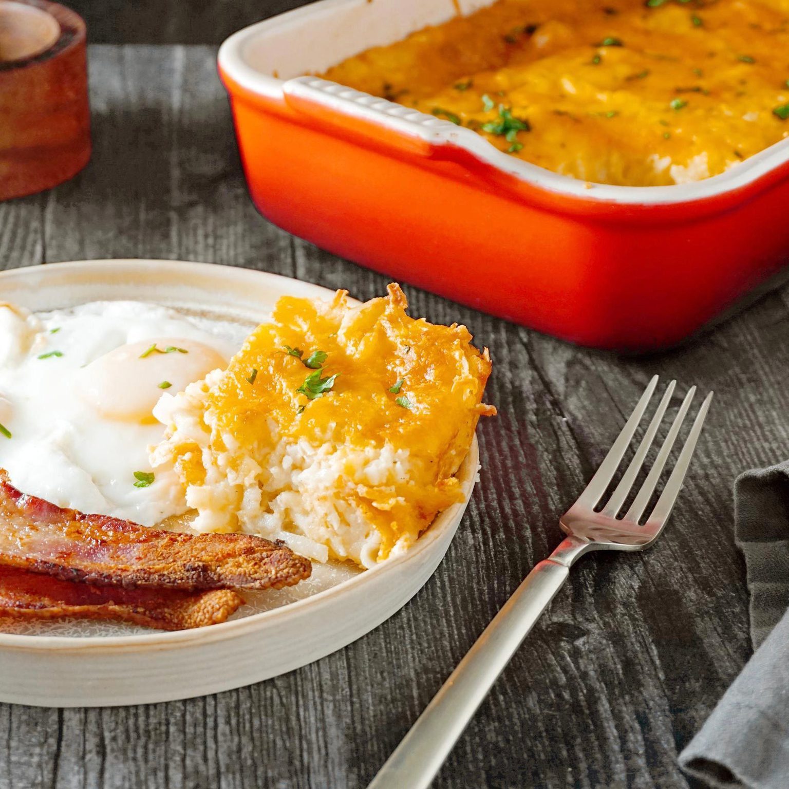 Breakfast plate holds eggs, bacon, and cheesy hash browns, resting on a dark wooden table beside a bright orange baking dish.