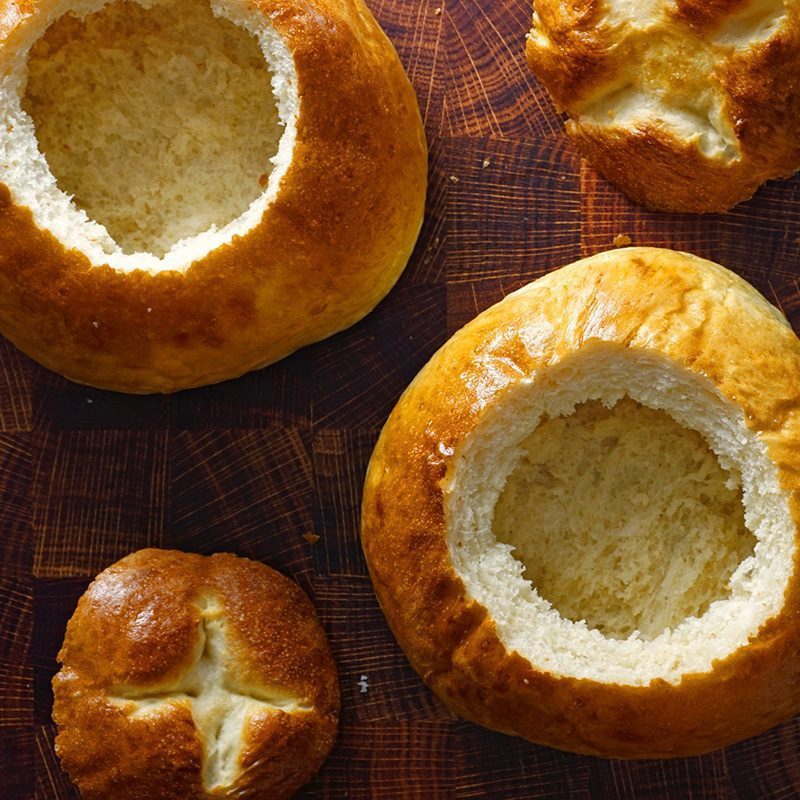 Hollowed bread bowls lie on a wooden surface, ready for filling, alongside whole bread rolls with scored tops.