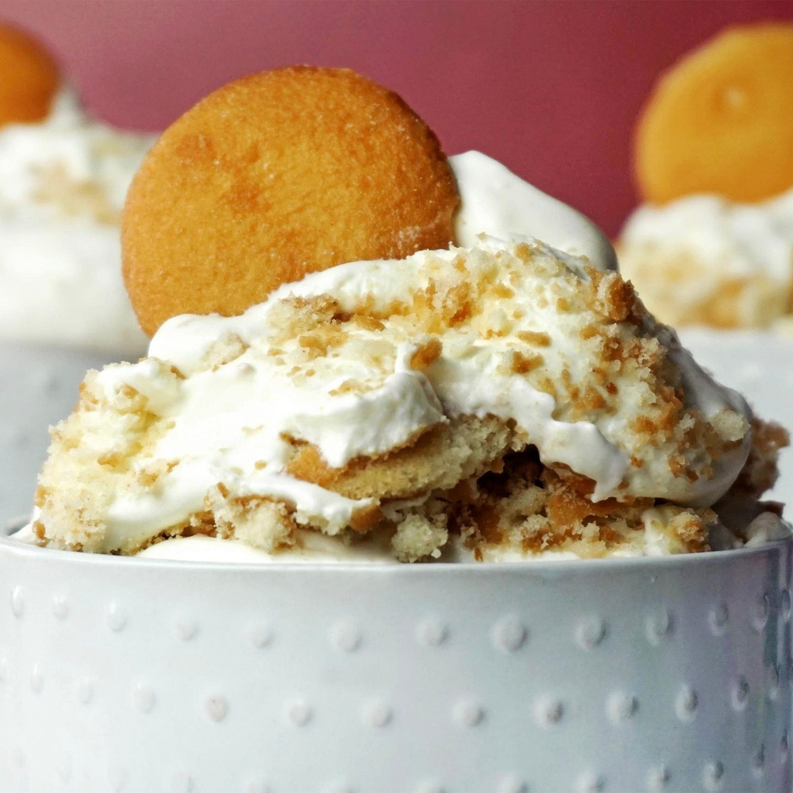 A dessert topped with crumbled cookies and a whole wafer rests in a textured white cup against a soft red background.