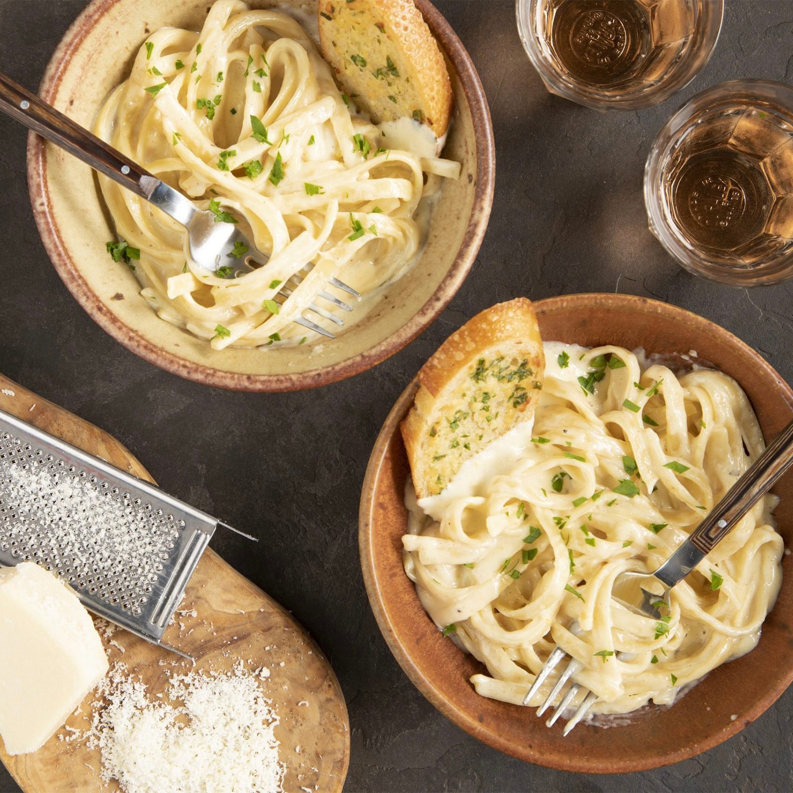 Two bowls of fettuccine alfredo with parsley and garlic bread, placed on a dark surface with grated cheese and two glasses nearby.