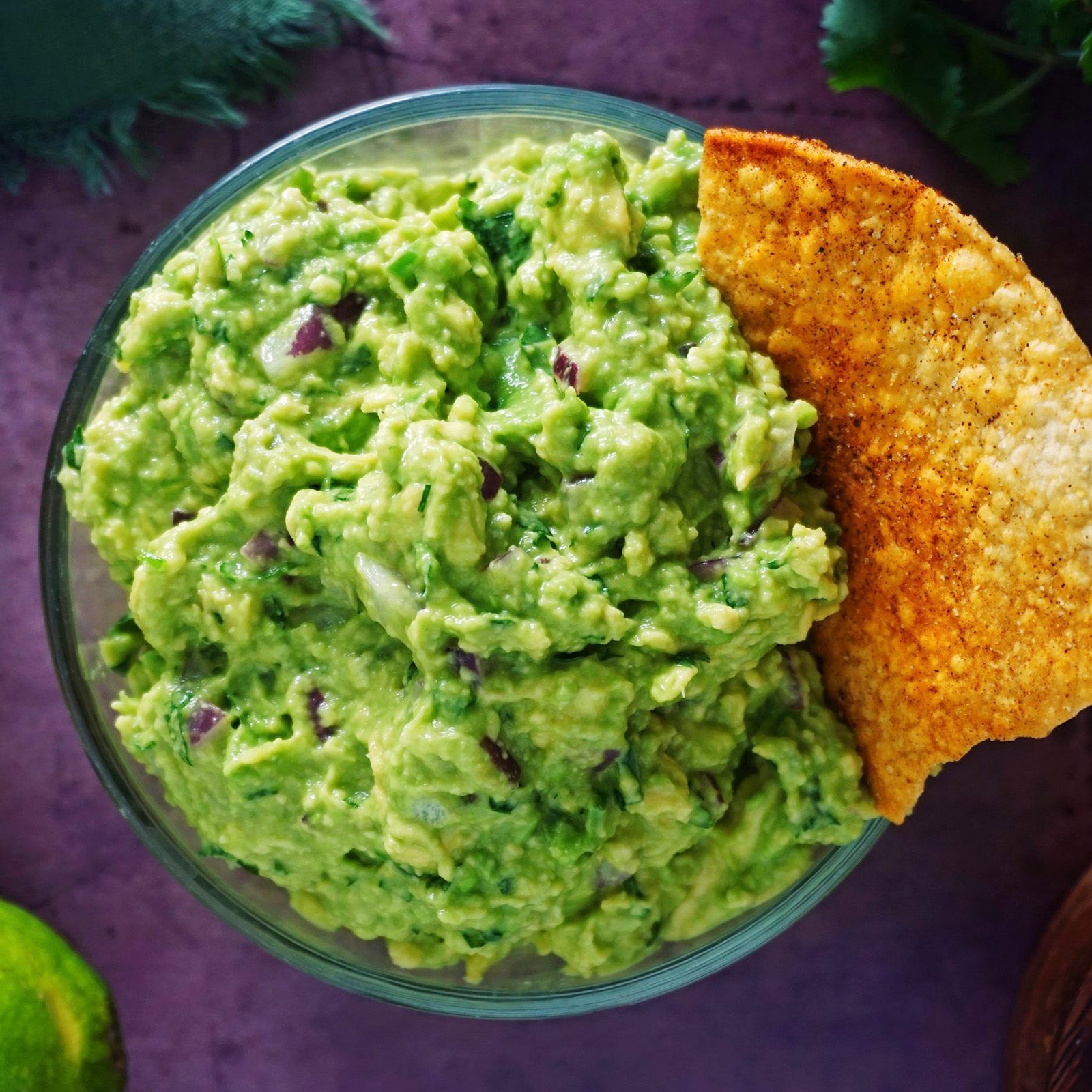 Guacamole sits in a glass bowl beside a tortilla chip; purple onion pieces are visible, surrounded by a dark surface with green accents.