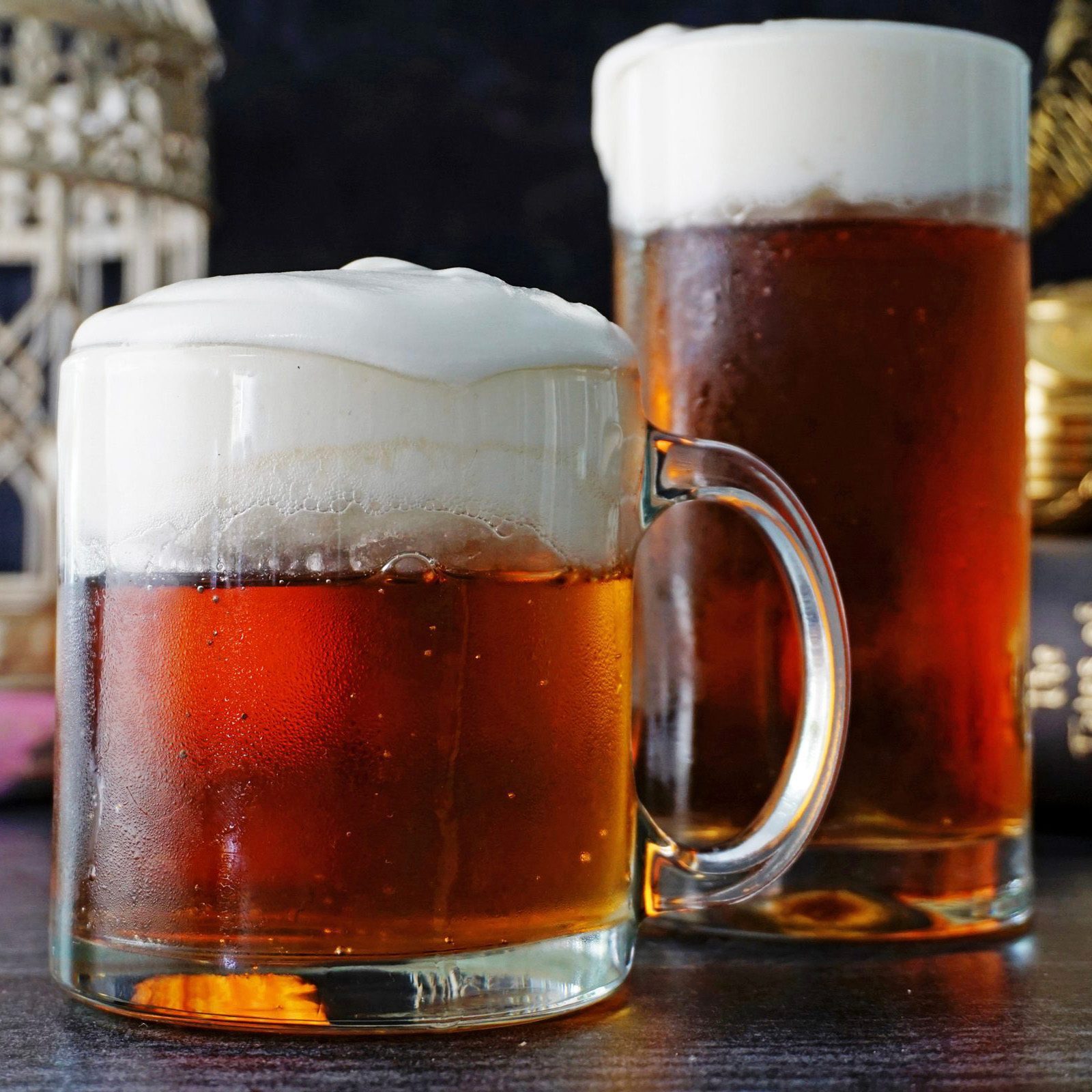 Frothy beer in glass mugs; foam topping amber liquid; set on a dark table with decorative objects in the background.