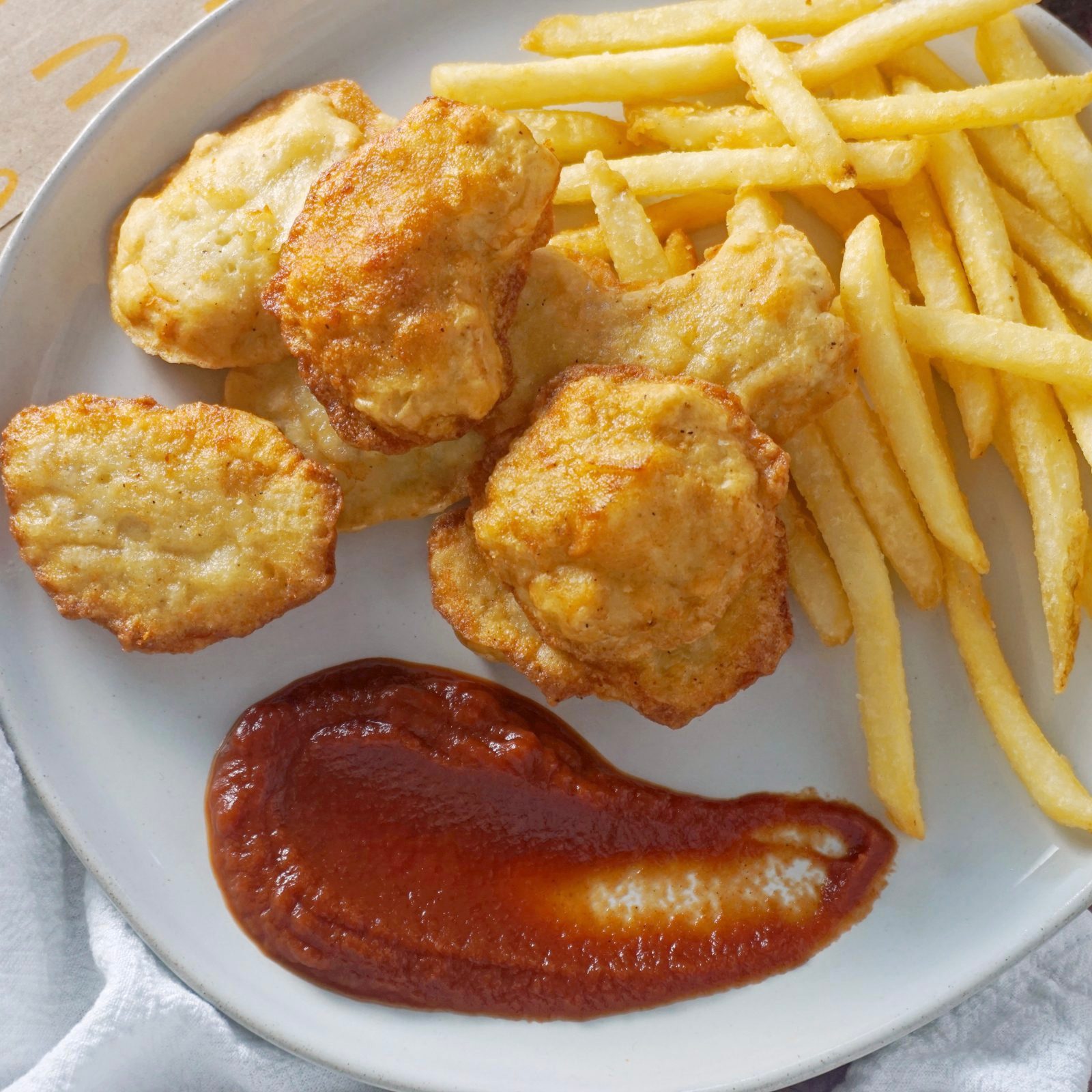 Chicken nuggets and fries rest on a plate with ketchup beside them. They are arranged neatly, suggesting a casual dining setting.