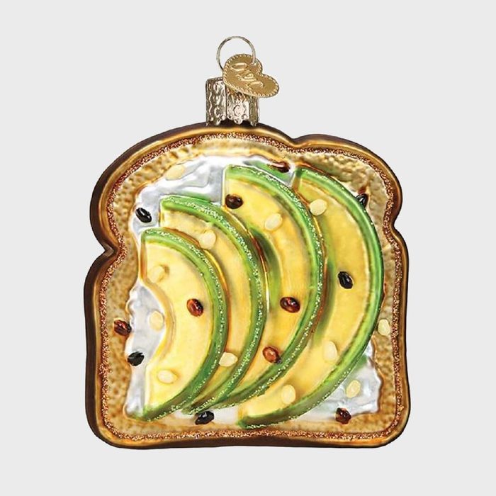 A toast ornament displays layered avocado slices with seeds, set against a plain background, topped with a decorative metal cap.
