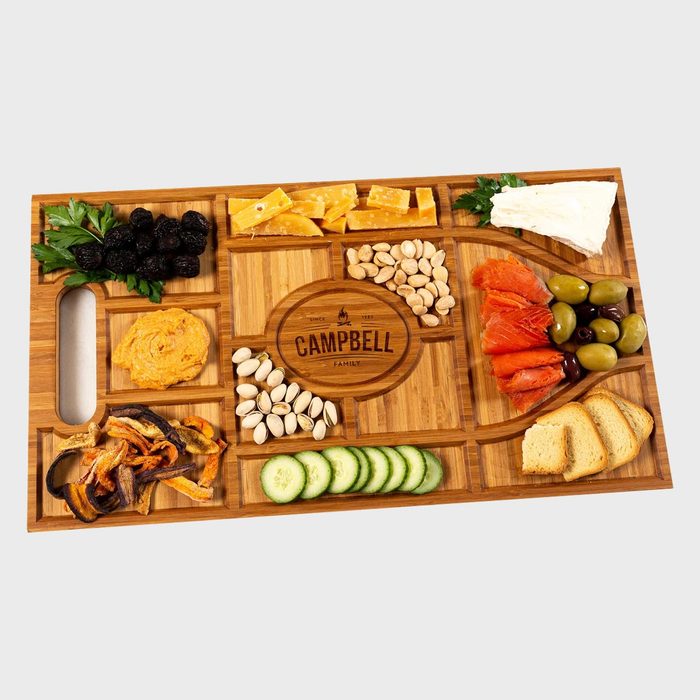 A wooden charcuterie board displays assorted cheeses, nuts, fruits, and vegetables. It features engraved text: 