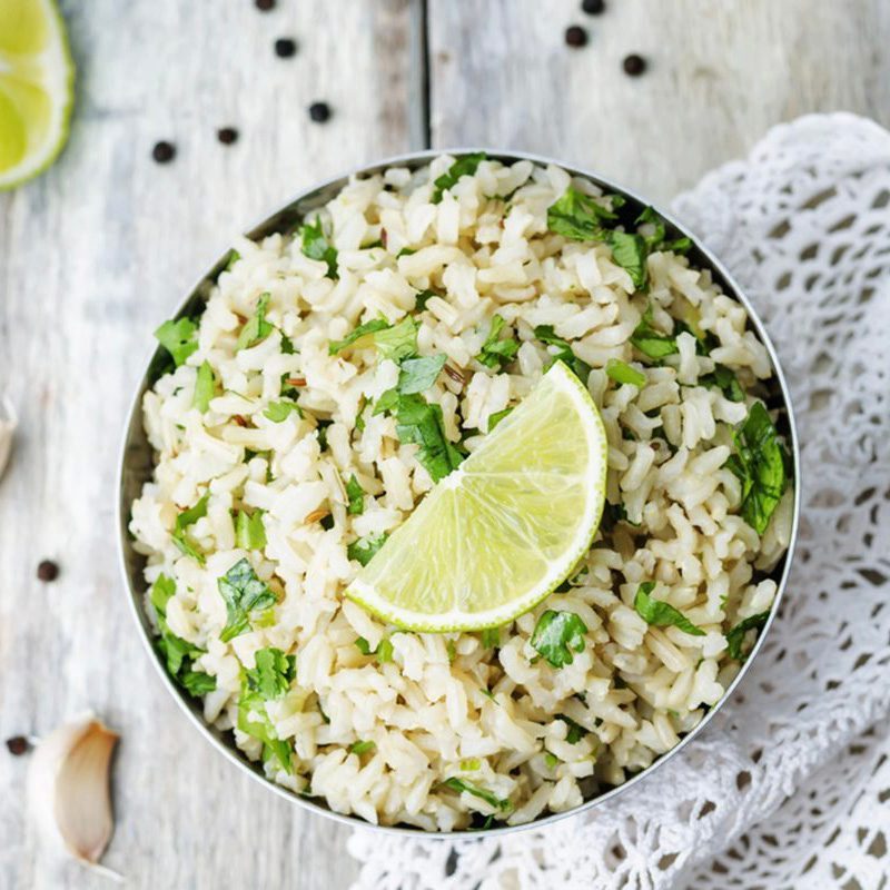 A bowl of rice garnished with cilantro and lime sits on a wooden surface with scattered peppercorns and garlic, near a lace doily.