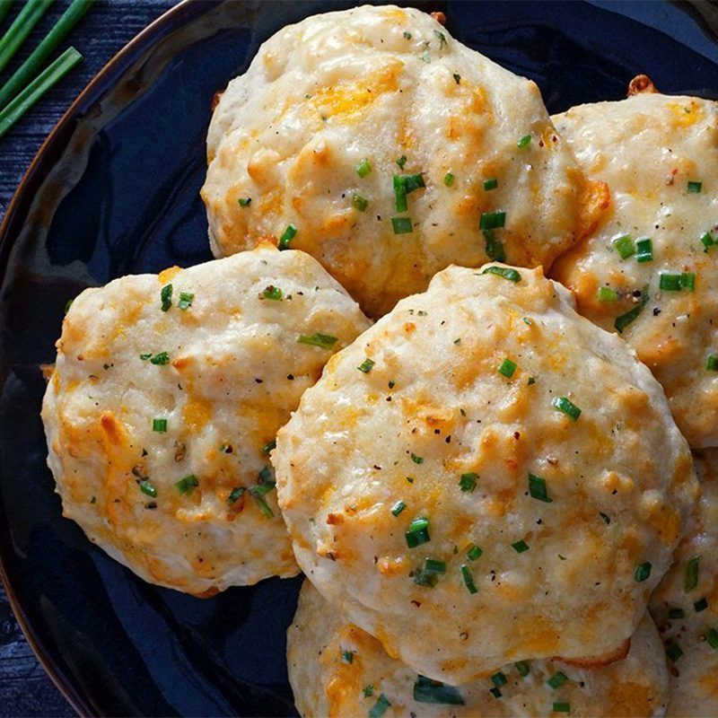 Biscuits sit on a dark plate, topped with melted cheese and chopped chives, surrounded by a few fresh chive stalks on a textured surface.